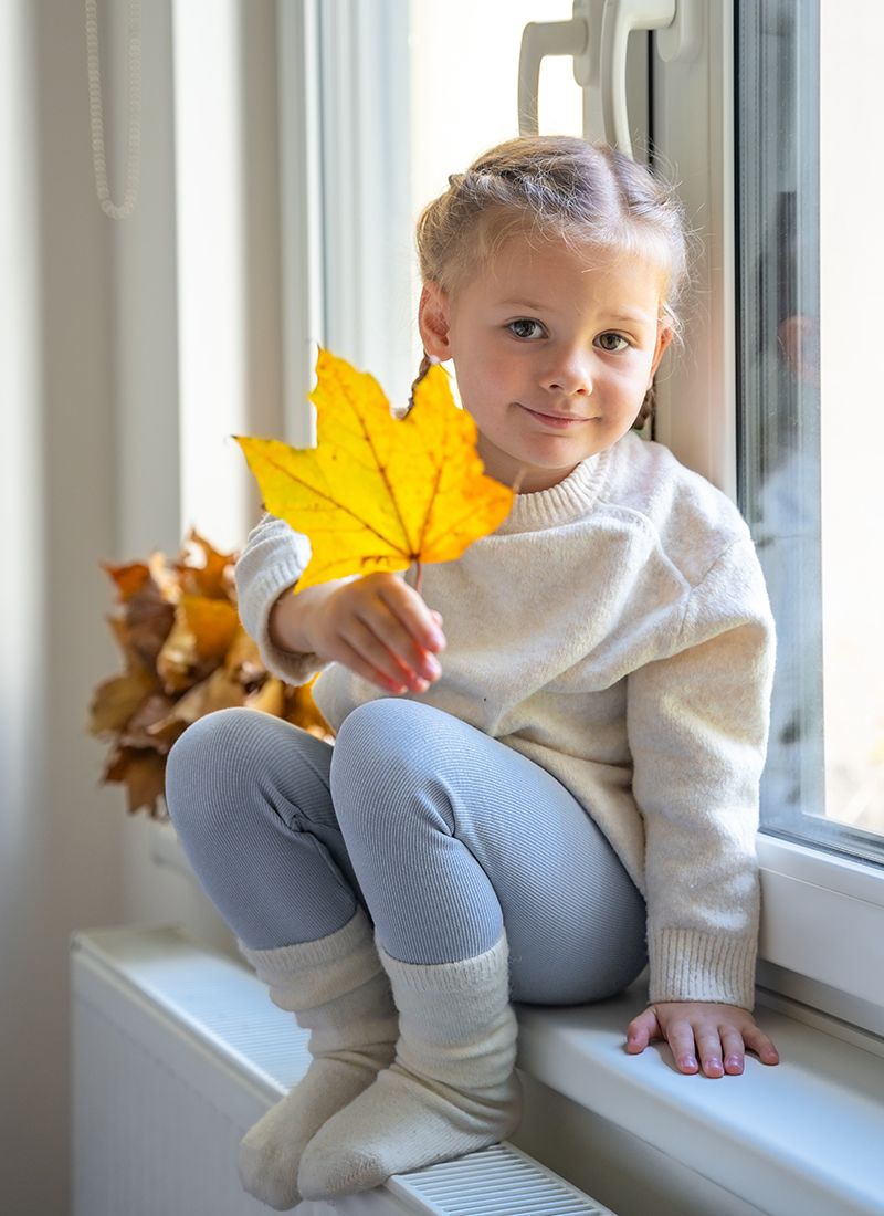 Una niña sentada en la ventana y con los pies en el radiador y una hoja de un árbol en la mano.