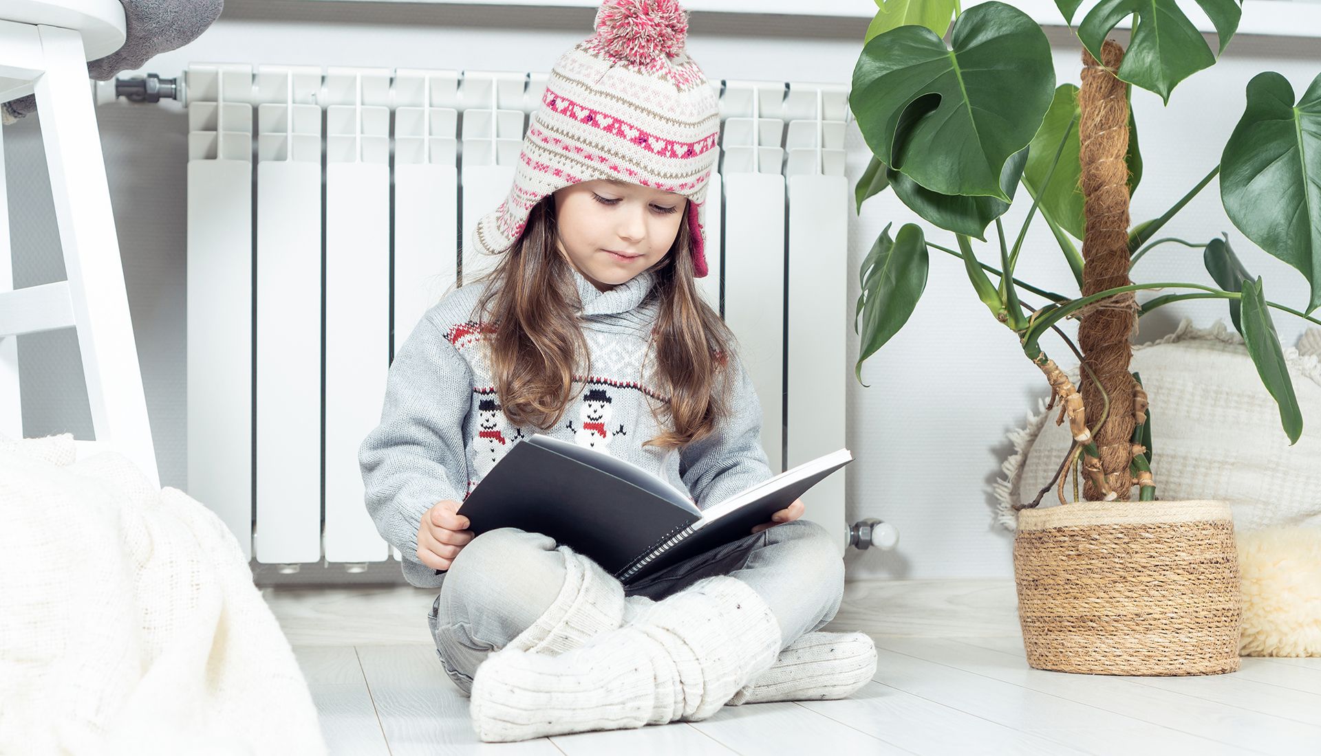 Niña sentada en el suelo al lado de un radiador. Ella está leyendo un cuaderno. La imagen hace referencia a la limpieza de calderas de gasóleo.